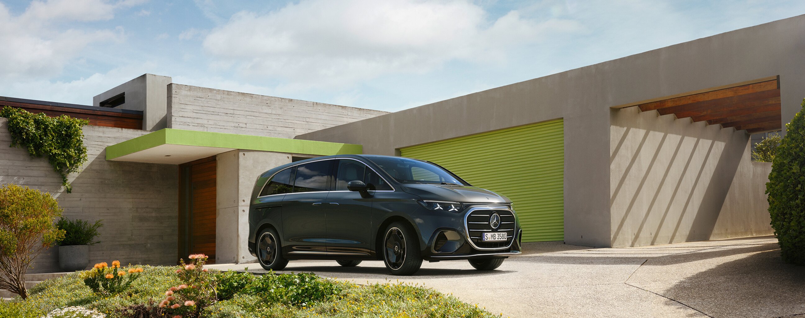 Das Bild zeigt einen Mercedes-Benz VLE, der vor einer modernen, architektonisch ansprechenden Villa geparkt ist. Die Fassade kombiniert Beton- und Glasstrukturen mit kreativen grünen Akzenten. Im Vordergrund befindet sich ein gepflegter Garten mit üppiger Vegetation und farbenfrohen Blumen, die der Szene eine lebendige und harmonische Atmosphäre verleihen. Der blaue Himmel und die natürlichen Elemente vermitteln eine ruhige und stilvolle Umgebung.
